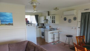 Een woonkamer met een bruine bank, een eethoek met een houten tafel en stoelen, een keuken met witte kastjes en een koelkast, en een plafondventilator.
