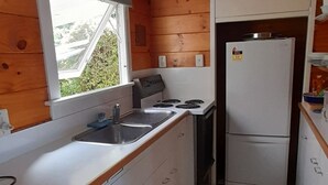 A compact kitchen with wooden walls, a window, a sink, a stove, and a refrigerator. - Quaint Lockwood River Cottage (Turangi)
