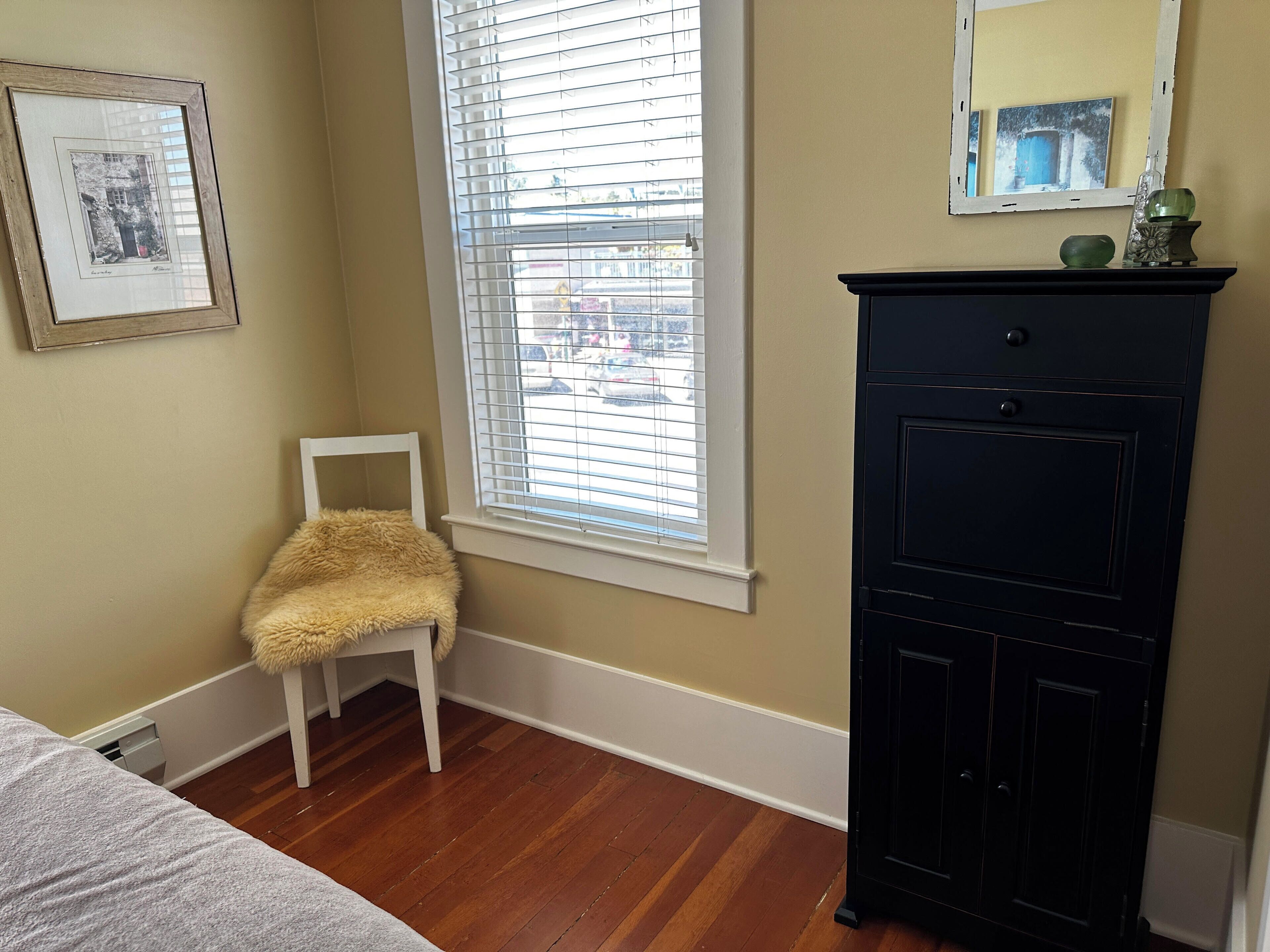 A room with a wooden floor, a white chair, a black cabinet, and a window with blinds.