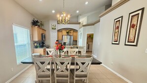 A dining area with a chandelier, a kitchen in the background, and framed artwork on the wall.
