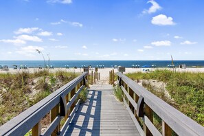 Ein Holzsteg führt zu einem Strand mit Sonnenschirmen und Liegestühlen.