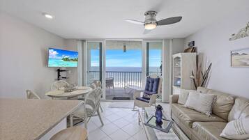 A living room with a TV, dining table, chairs, a sofa, and a balcony with a view of the ocean.