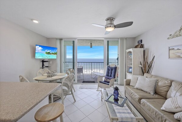 A living room with a TV, dining table, chairs, a sofa, and a balcony with a view of the ocean.