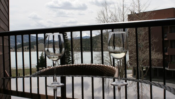 Un balcón con dos copas de vino y una silla de mimbre, con vistas al agua y montañas.