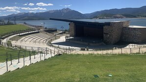 Un teatro con escenario y zona de asientos, rodeado de césped y un cuerpo de agua con montañas al fondo.