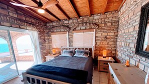 Un dormitorio con pared de piedra, cama con cabecera de madera, dos mesitas de noche con lámparas y vistas a través de un ventanal.