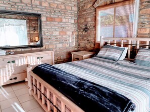 A bedroom with a brick wall, a bed with striped bedding, a dresser with drawers, a mirror, and a window with a view.