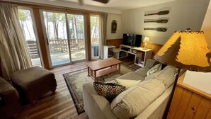 Sala de estar com sofá, mesa de centro, TV e ventilador de teto. Há uma porta de vidro deslizante que dá para um deck externo.