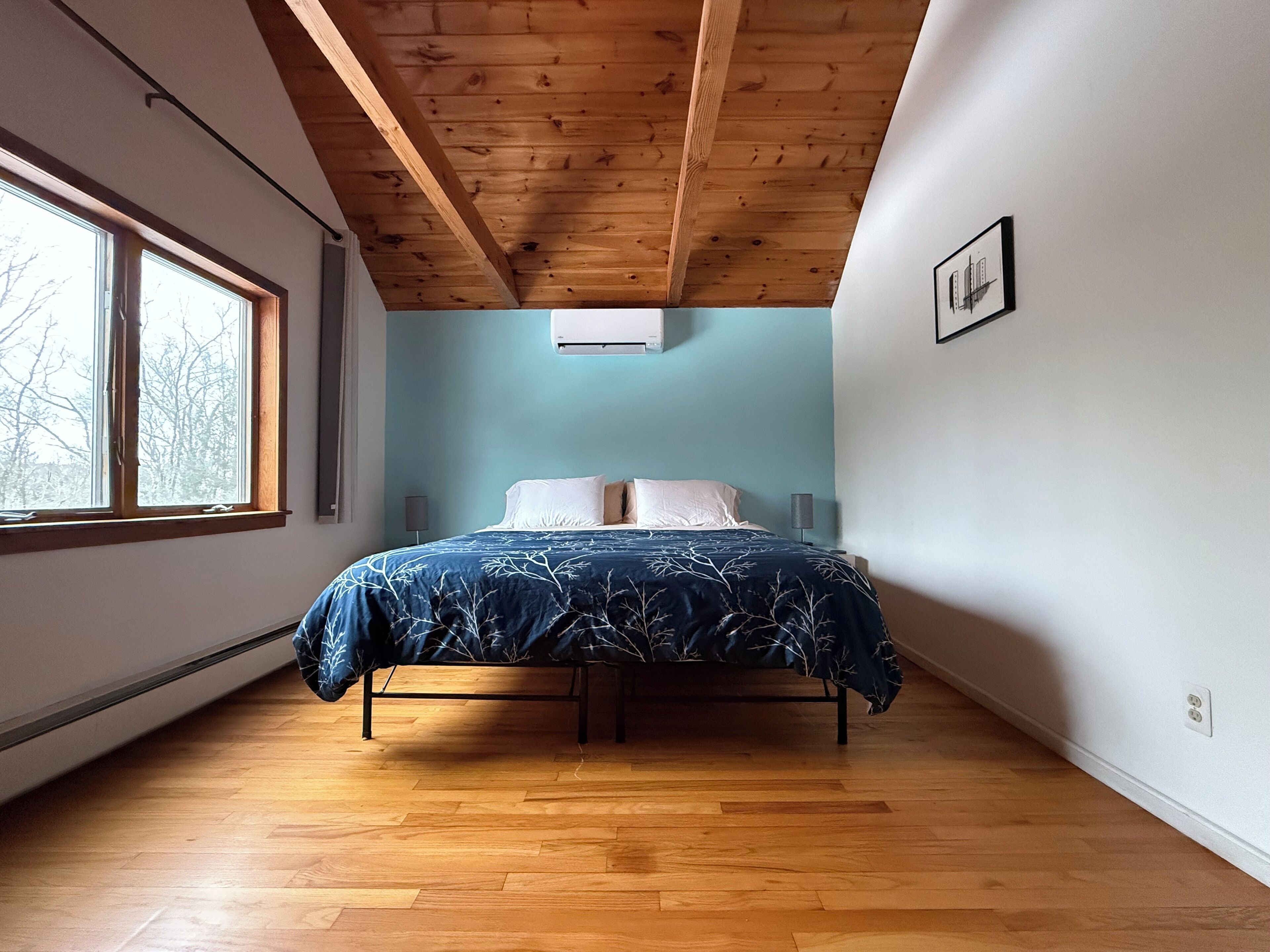 Een slaapkamer met een houten plafond, een bed met een blauw bloemenbedekking, een raam met uitzicht op bomen en een wandairconditioner.