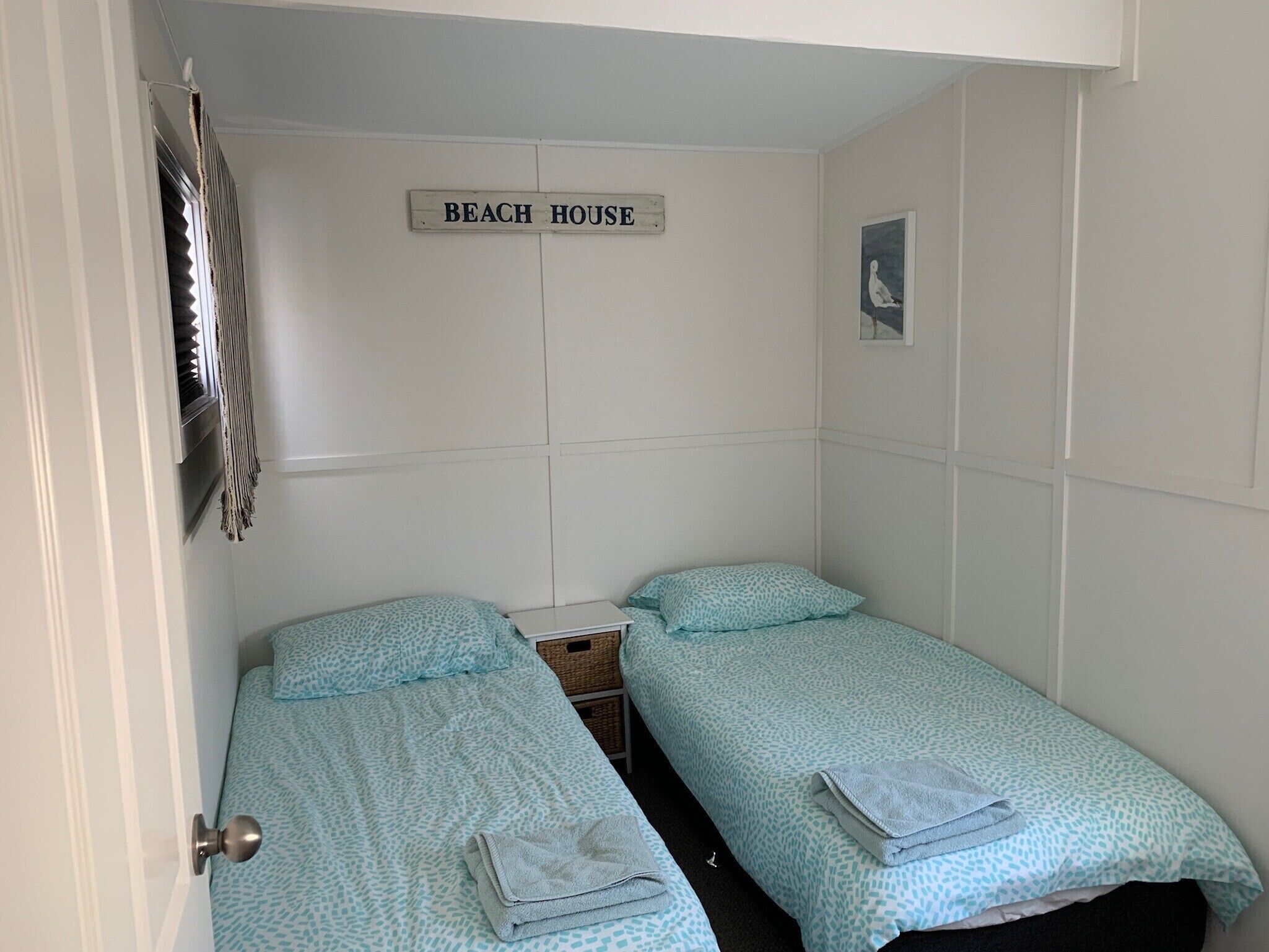 Una stanza piccola e pulita con due letti, un cartello con la scritta 'BEACH HOUSE' e un quadro appeso al muro.
