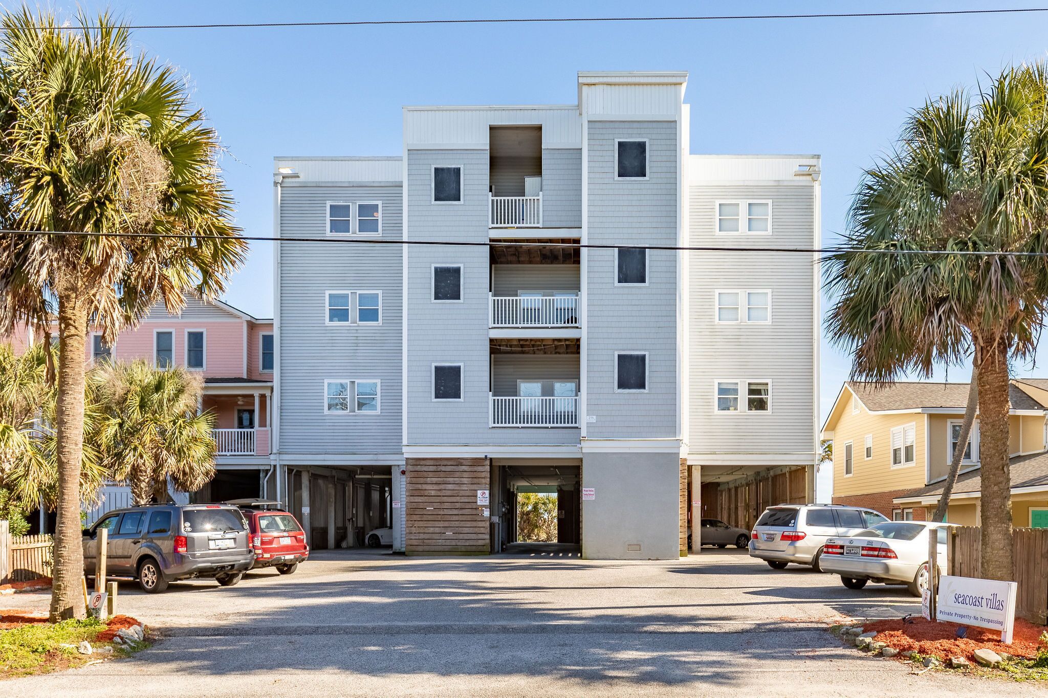 A multi-story residential building with a central entrance, parking area, and surrounding palm trees.
