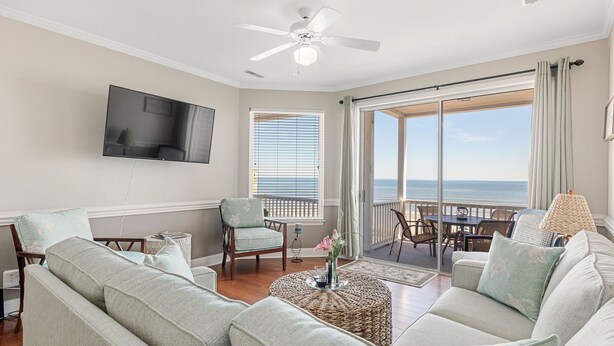 A living room with a large window, a ceiling fan, a flat-screen TV, and a balcony with a table and chairs. - Seacoast 7- Top Floor Villa - Unobstructed Ocean Views - Elevator (Folly Beach)