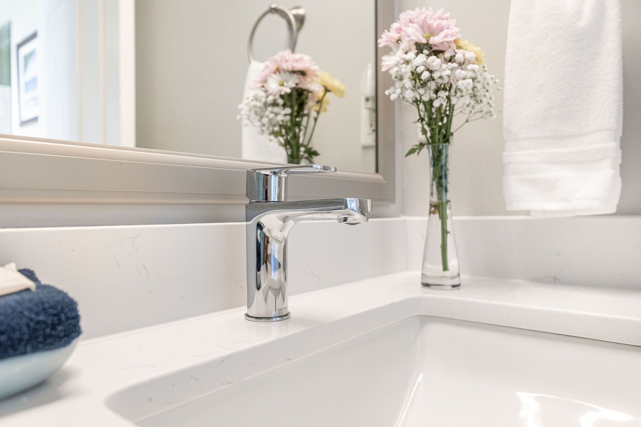 A bathroom with a chrome faucet, a white sink, and a mirror reflecting flowers and towels.