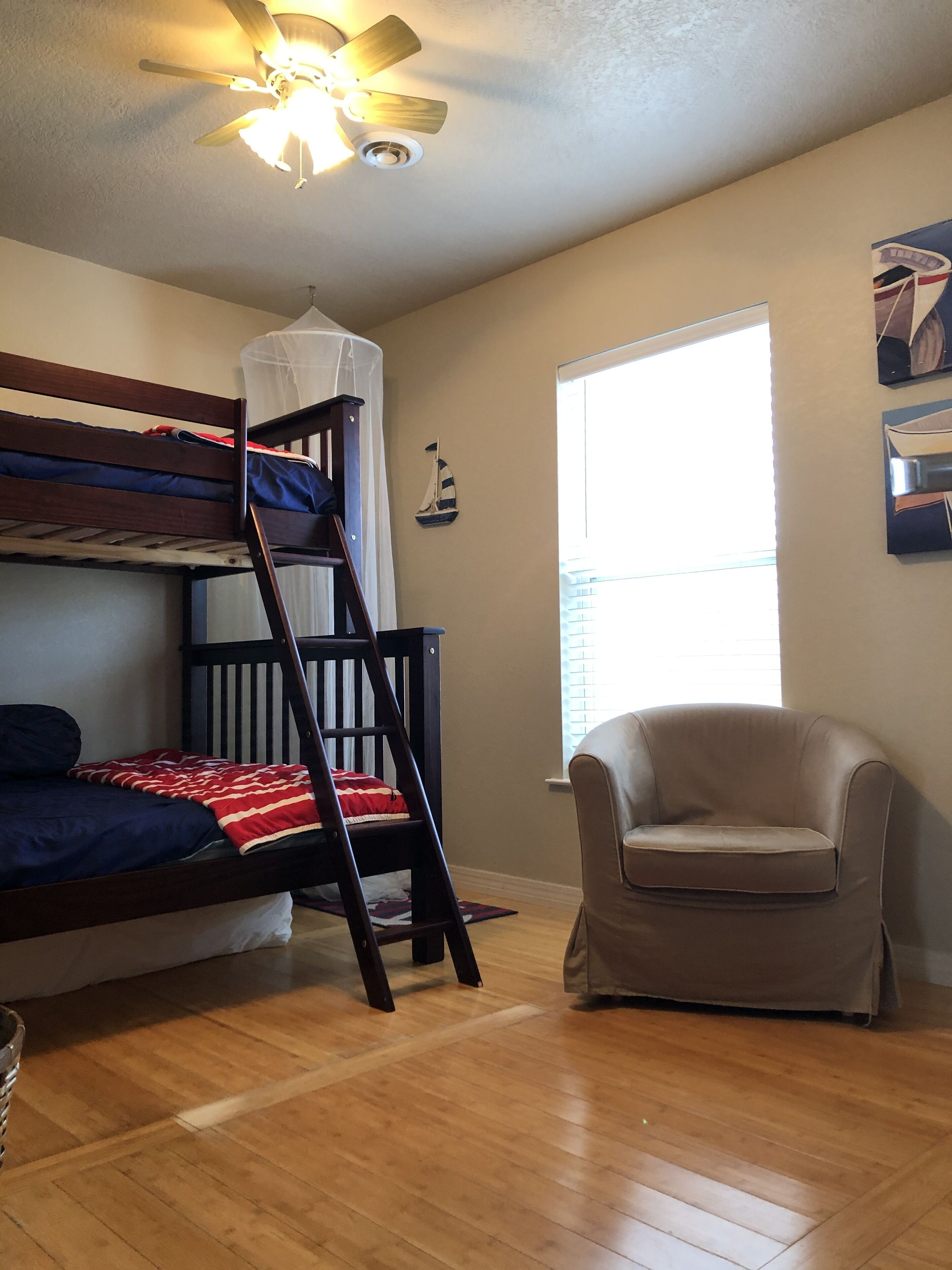 A room with a bunk bed, a ladder, a chair, a ceiling fan, and a window.
