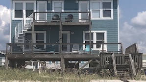 A two-story beach house with a wooden deck, multiple windows, and a staircase leading to the upper floor.