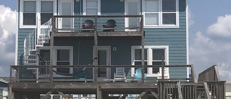 Een tweelaags strandhuis met een houten terras, meerdere ramen en een trap naar de bovenverdieping.