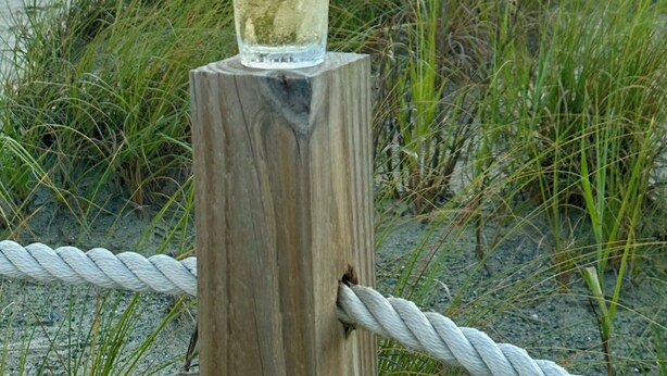 A glass of white liquid on a wooden post by the beach with grass and the ocean in the background.