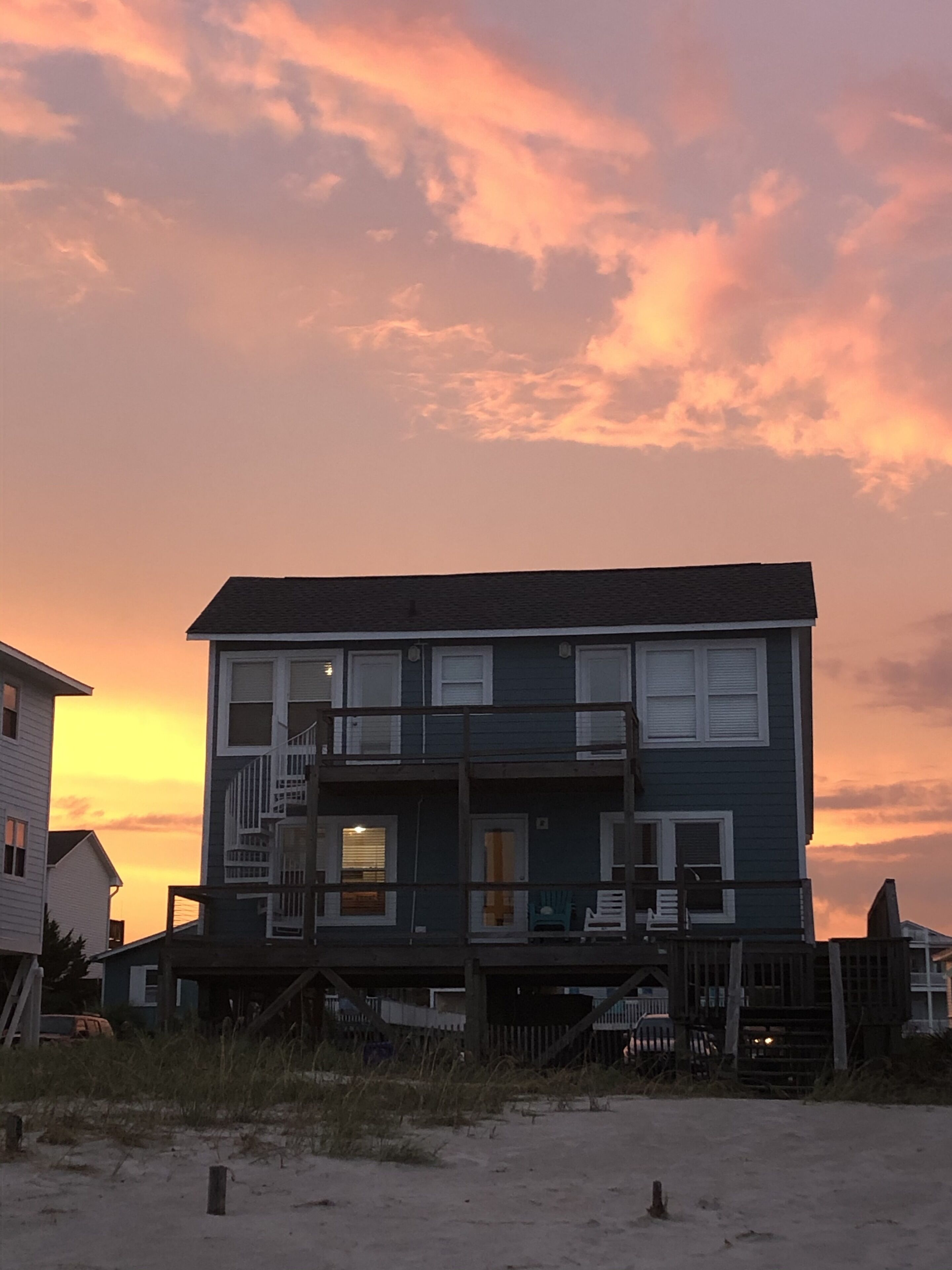 Une maison de plage à deux étages, dotée d’un balcon, située sur une plage de sable avec un coucher de soleil en toile de fond.