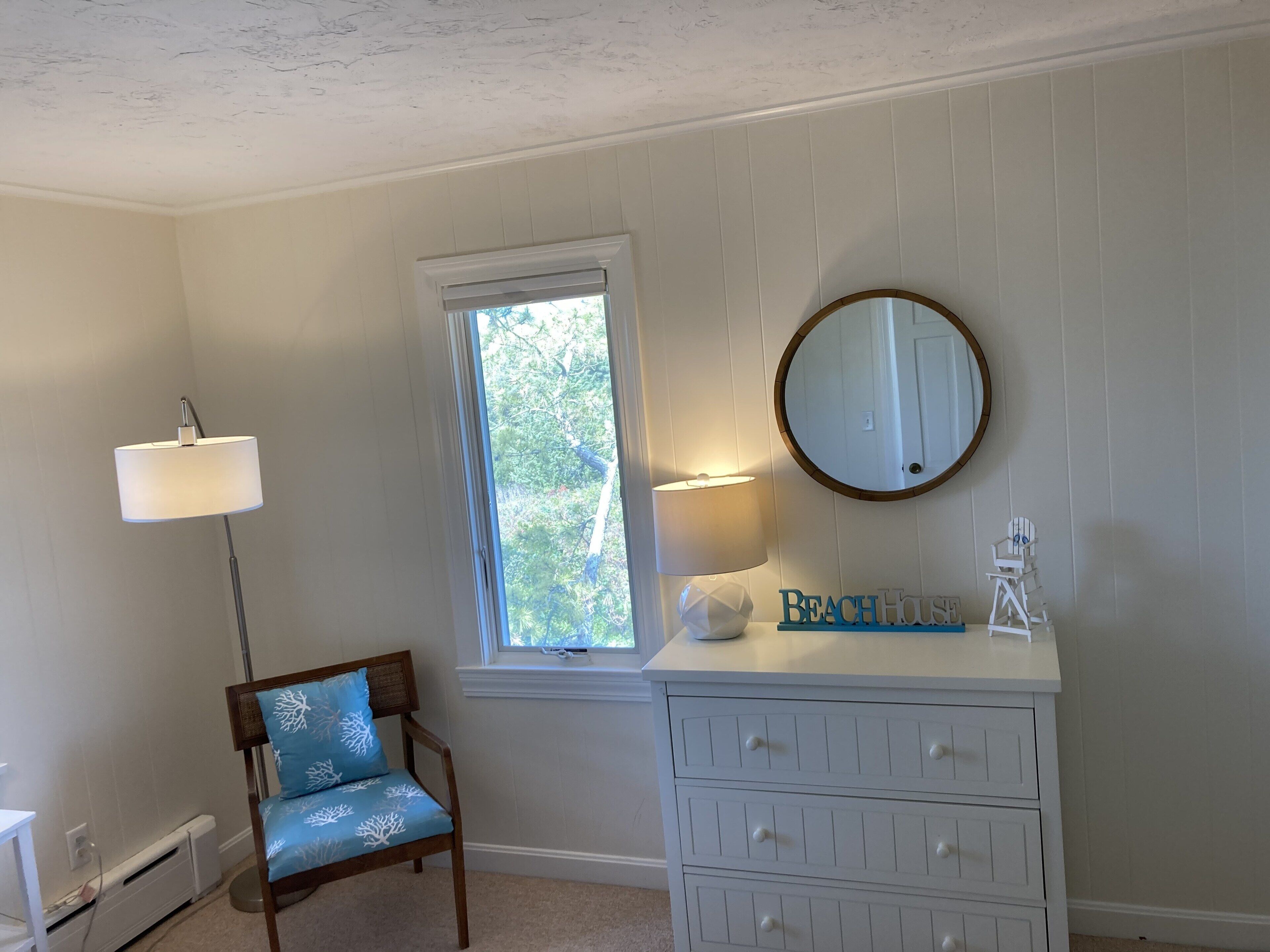 A room with a window, a chair with a blue cushion, a dresser with drawers, a lamp, and a mirror.