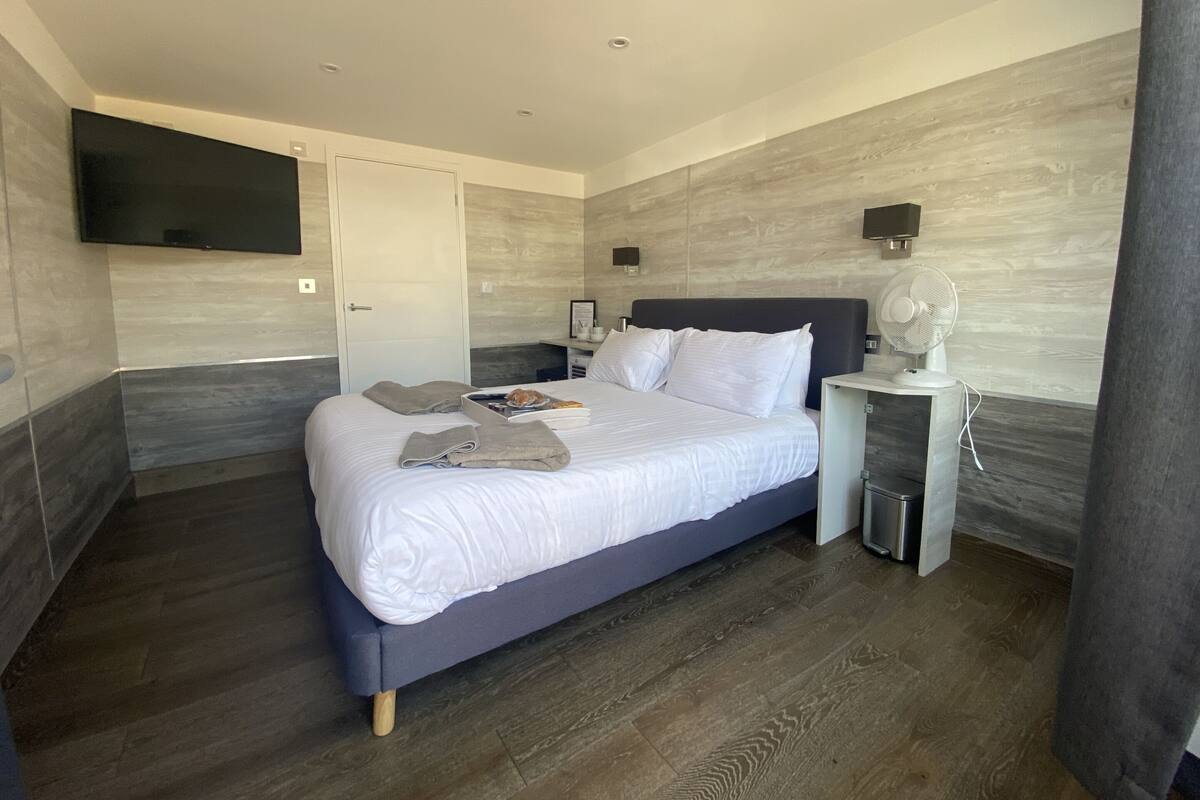 A modern bedroom with a large bed, a flat-screen TV mounted on the wall, a bedside table with a lamp, and a small waste bin.