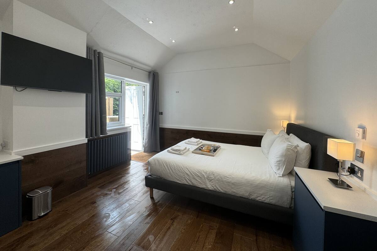 A modern hotel room with a large bed, a bedside table with a lamp, a built-in TV, and a view of the outdoors.