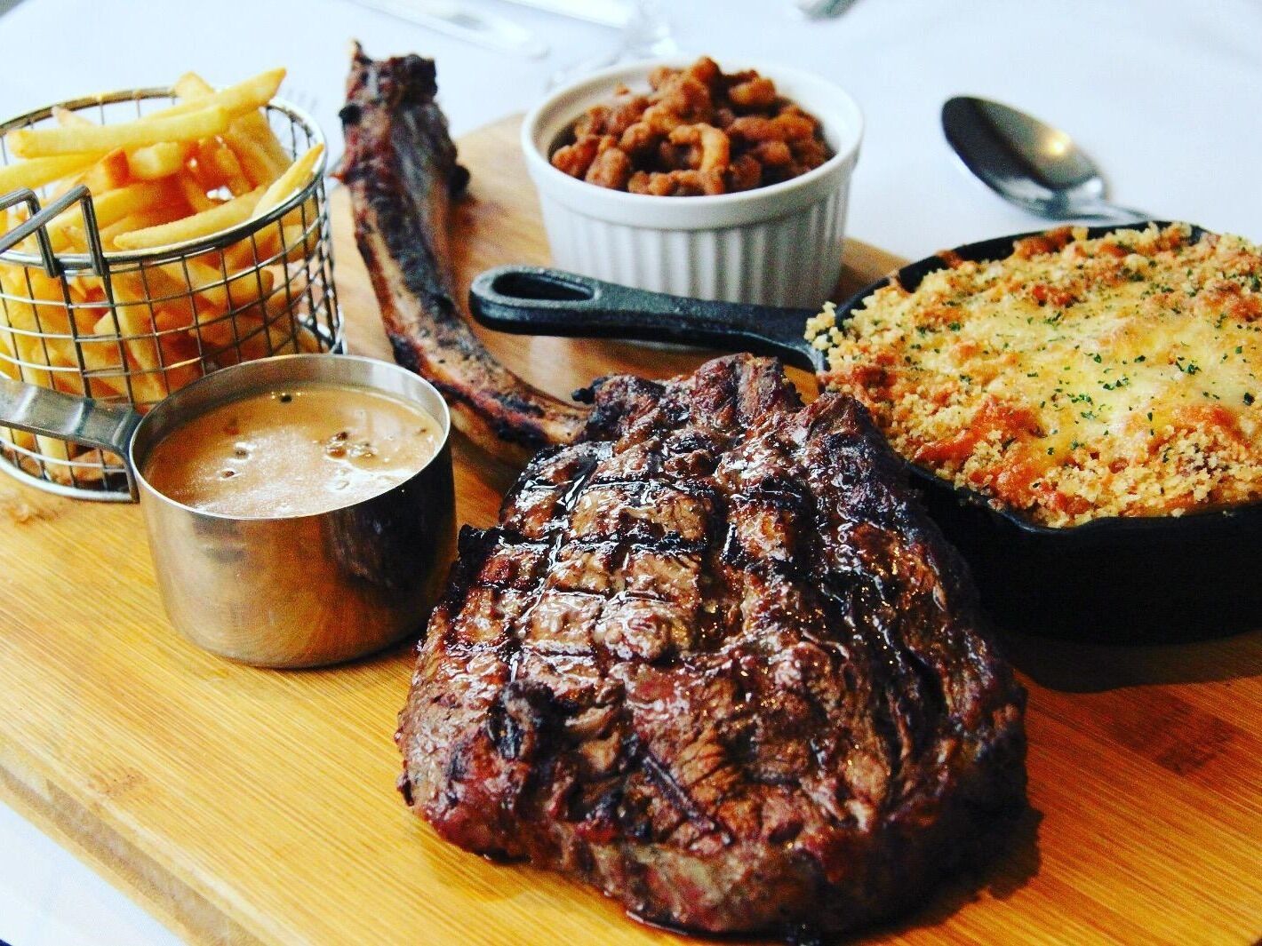 A steak with fries, beans, and a baked dish on a wooden board.