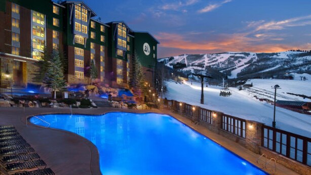 Un hotel con piscina, montañas nevadas al fondo e iluminación exterior destacada.