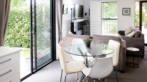 Un salon moderne avec une table à manger en verre, des chaises blanches, un canapé marron et un téléviseur posé sur un meuble.