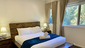 A neatly made bed with white linens, a blue throw blanket, and a rolled towel. There are two bedside tables with lamps and a clock. The room has a wooden headboard and a window with curtains. - Olinda Two Bedroom Apartment B2 - This beautiful and comfortably furnished apartment is centrally located in the village of Olinda. (Olinda)