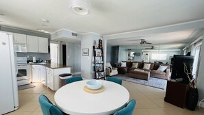 Una cocina moderna con armarios blancos, un comedor con sillas color turquesa y una sala de estar con sofá marrón y televisor.