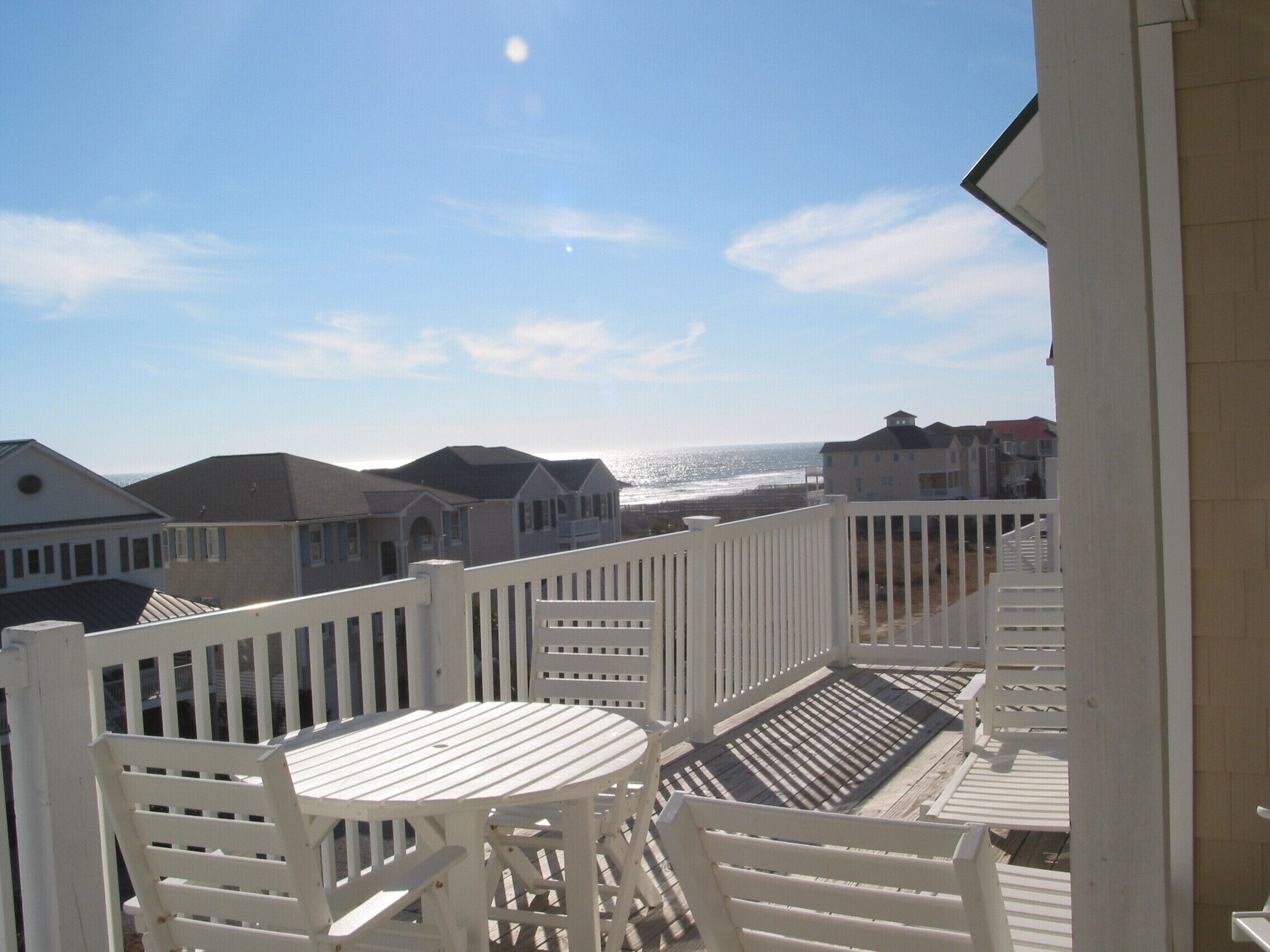 A balcony with white railings, a table, and chairs, overlooking a beach with houses in the background.
