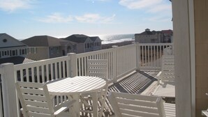 Un balcon avec des balustrades blanches, une table et des chaises, donnant sur une plage avec des maisons en arrière-plan.
