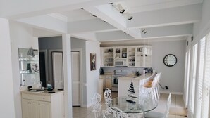 Une cuisine moderne avec des meubles blancs, un coin repas doté d’une table en verre et de chaises blanches, et une grande horloge au mur.