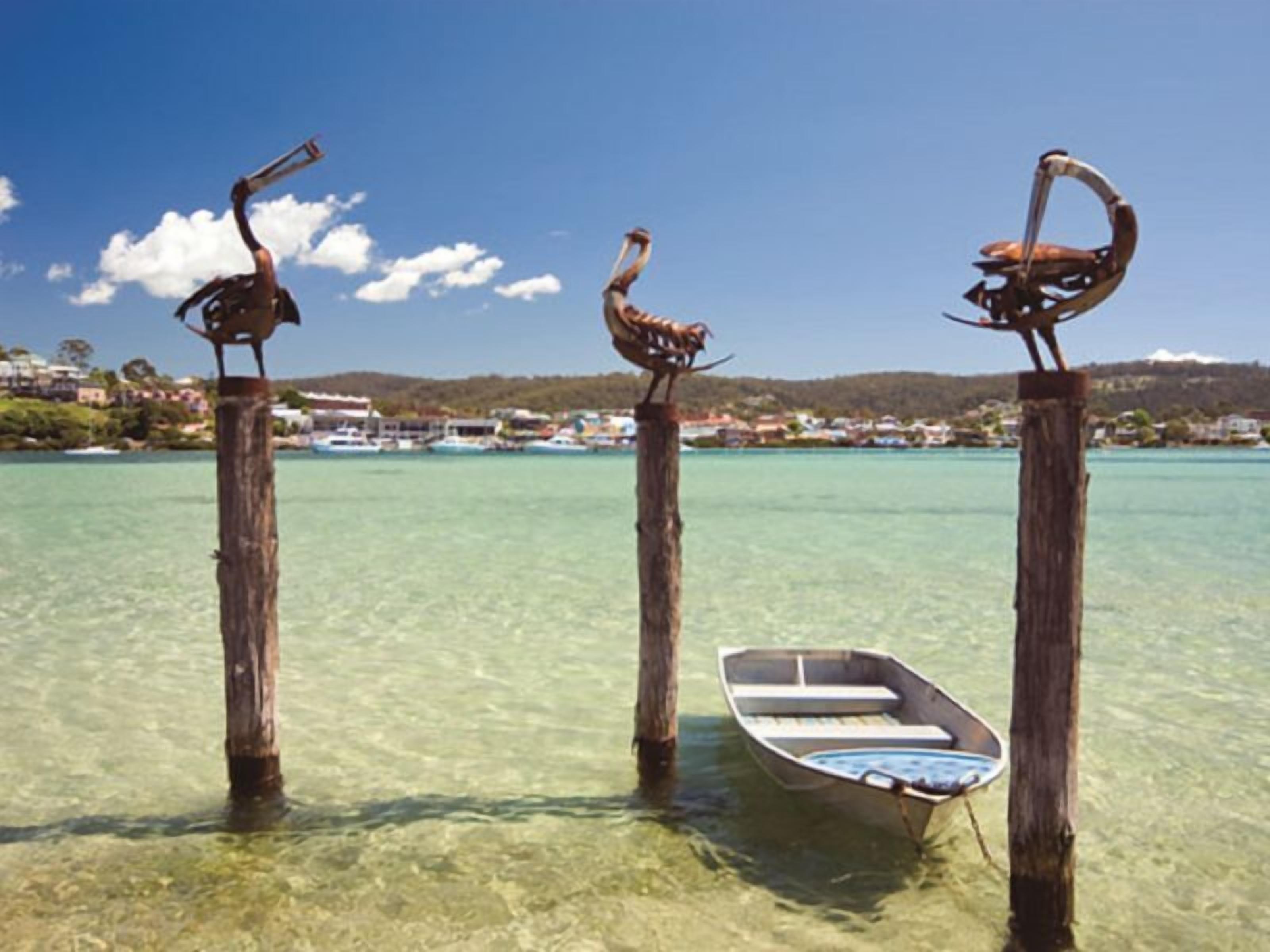 Tres postes de madera con esculturas de aves en agua clara y poco profunda, una lancha atada a uno de los postes y un pueblo costero al fondo.