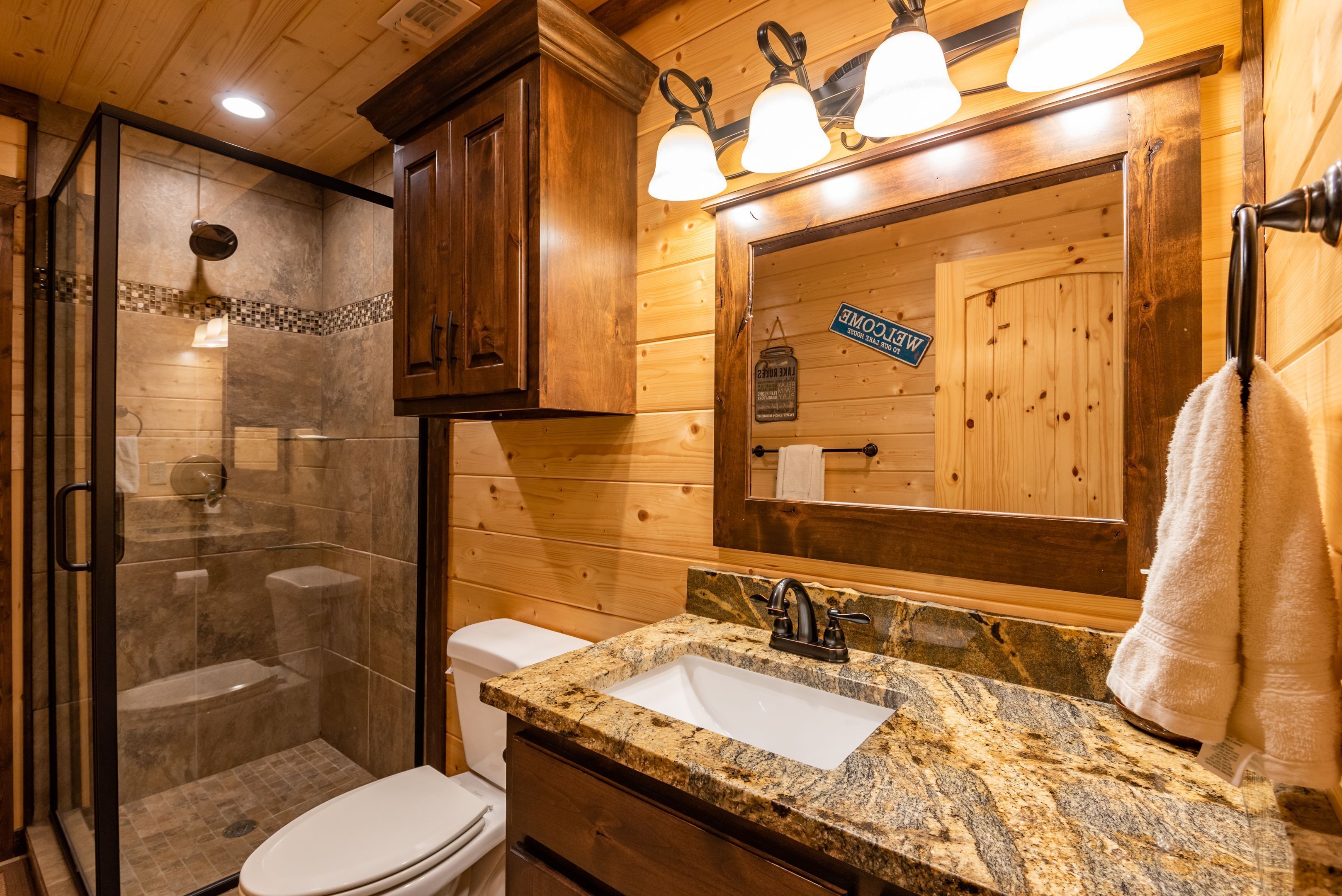 Baño de cabaña de madera con ducha acristalada, inodoro blanco y lavamanos con encimera de granito.