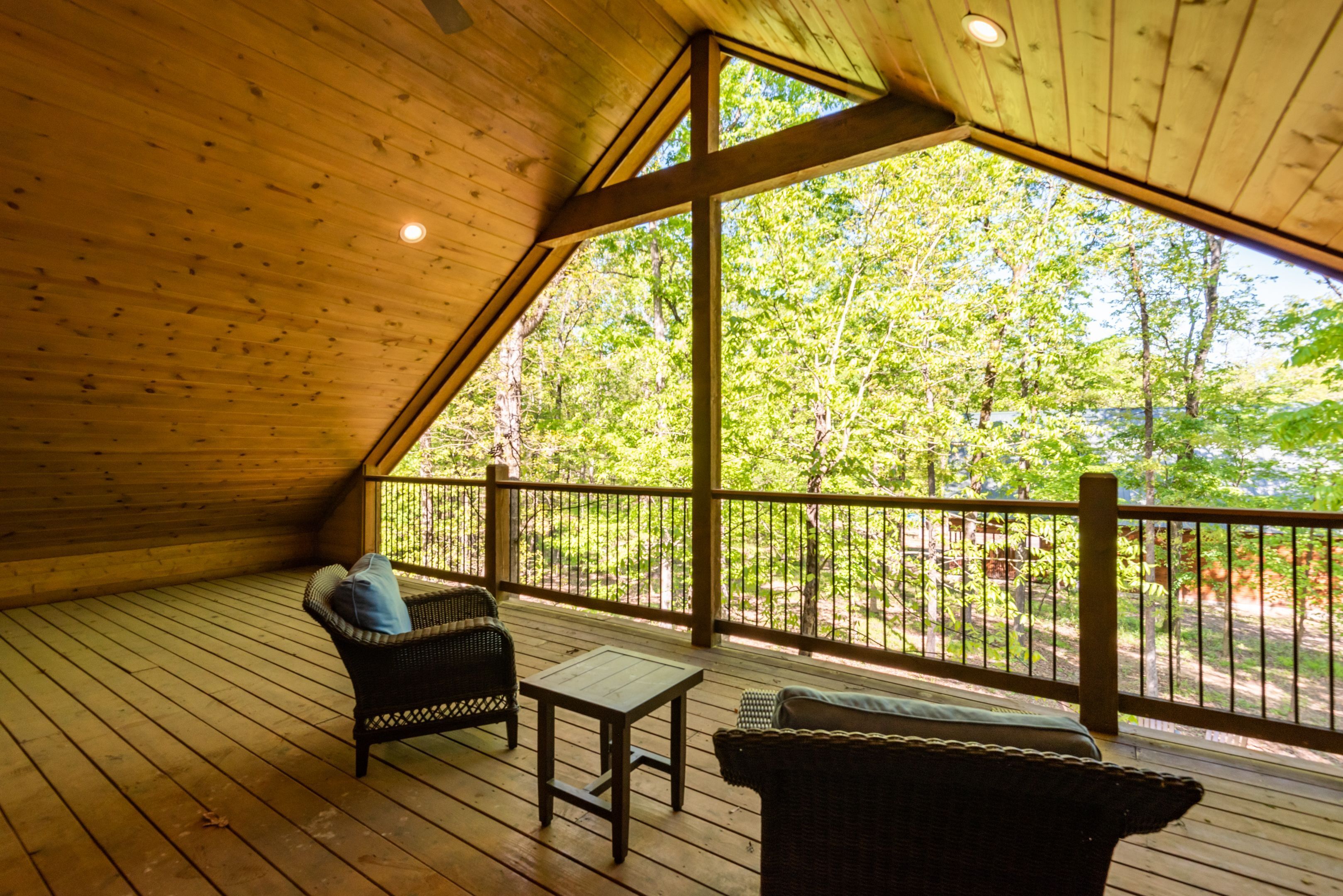 A wooden deck with a high ceiling and large windows, overlooking a forested area.