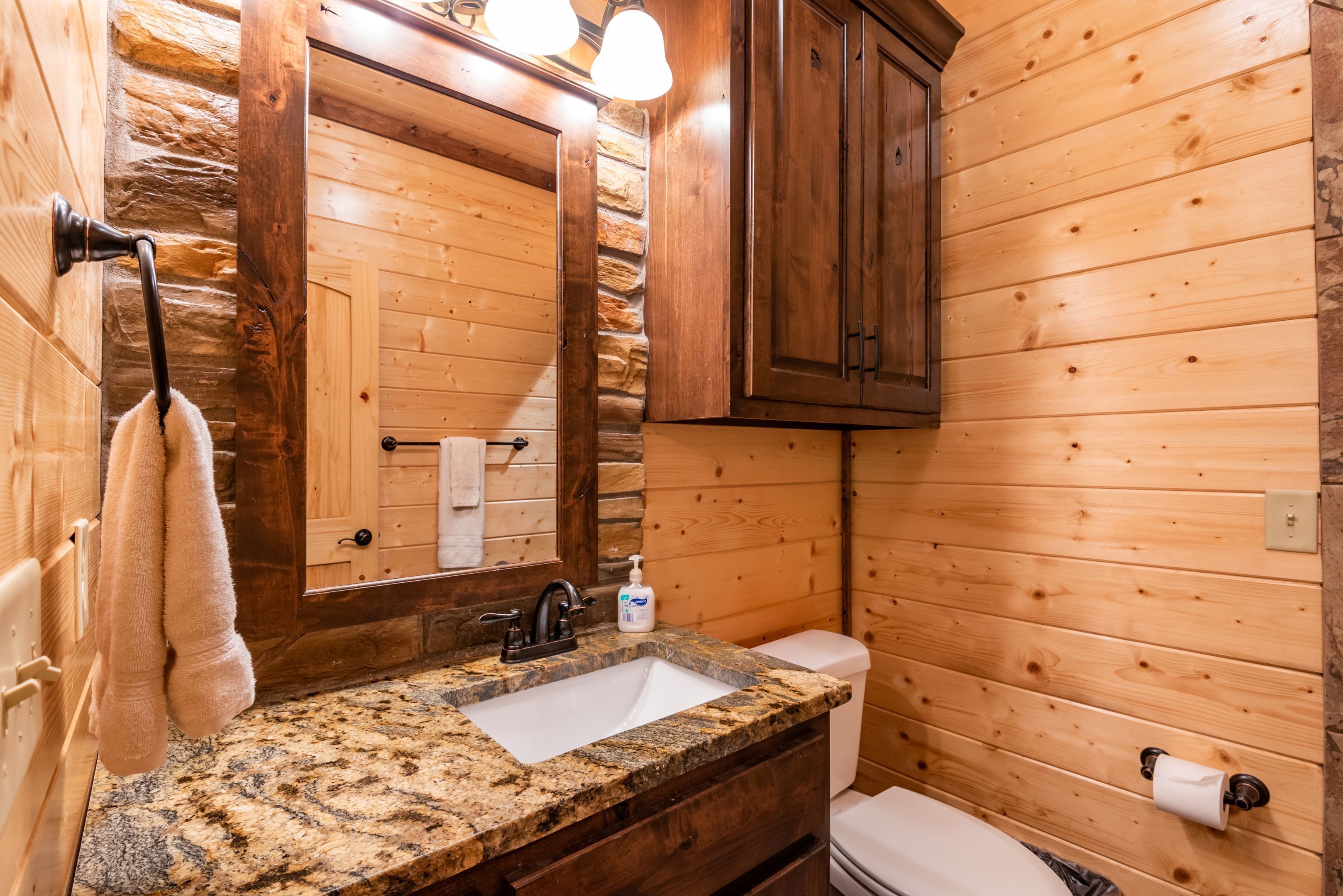 A bathroom with a granite countertop sink, a mirror, wooden cabinets, a towel hanging on the wall, and a toilet.