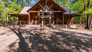 A large log cabin with a central staircase, surrounded by a gravel driveway and a metal fence.