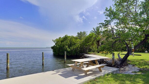 A lakeside picnic area with a wooden table and benches, surrounded by lush greenery and a clear view of the water. - Cottage with Pool and Dockage, just minutes to Key West (Cudjoe Key)