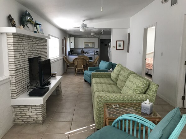 Una sala de estar con chimenea de ladrillo, televisor, sofá verde estampado y un comedor con sillas de mimbre.