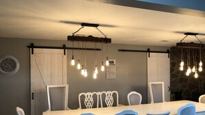 A dining area with a long table, blue upholstered chairs, hanging light fixtures, and a wall clock.