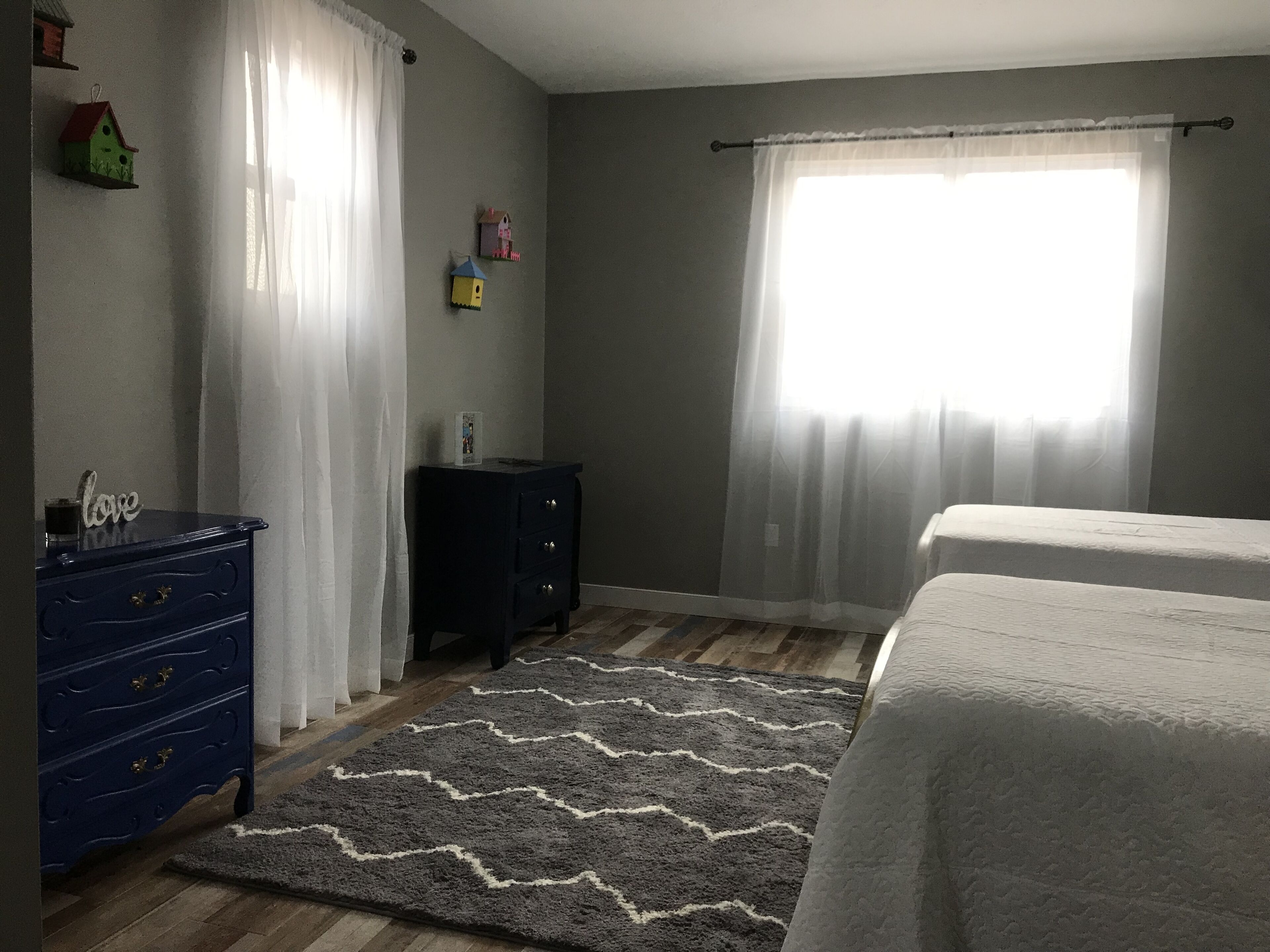 A bedroom with a bed, a dresser, a nightstand, a window with sheer curtains, and a wall-mounted birdhouse.