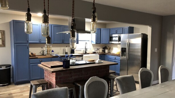 A modern kitchen with blue cabinets, a central island, hanging light fixtures, and a stainless steel refrigerator.