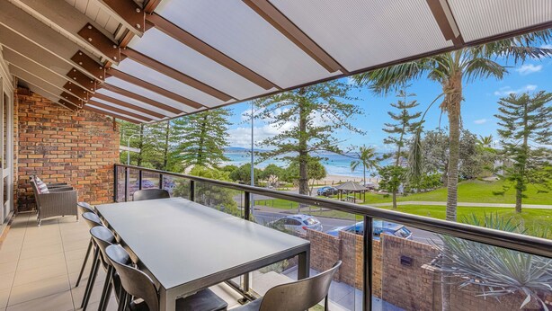 A balcony with a view of the beach, a table, chairs, and a brick wall.