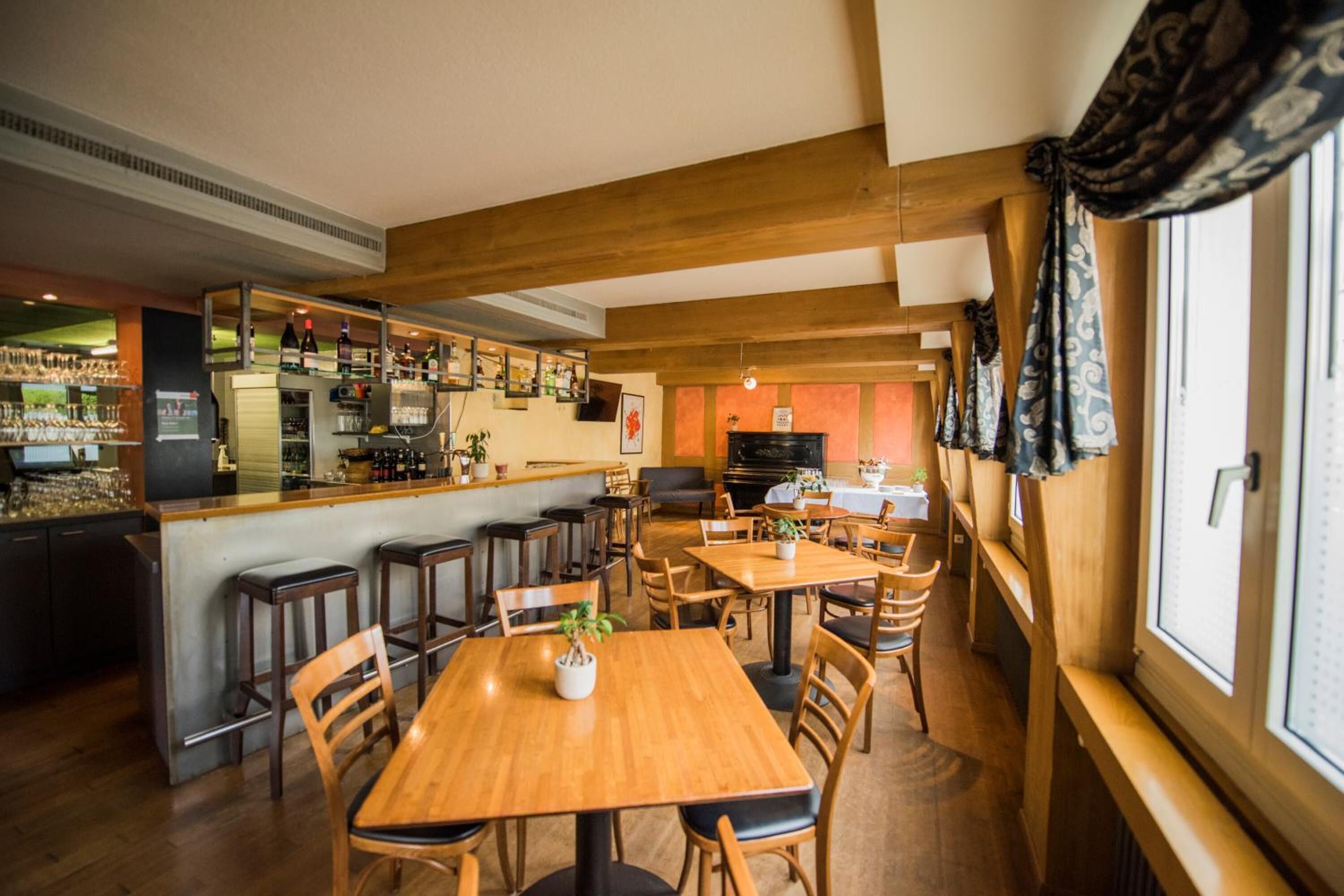 Un ristorante con tavoli e sedie in legno, un'area bar con scaffali pieni di bottiglie e un pianoforte sullo sfondo.
