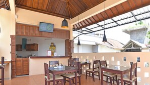 A dining area with wooden tables and chairs, a kitchen in the background, and a tiled floor. - RedDoorz @ Uluwatu Bali (Pecatu)