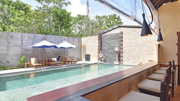 A pool area with a wooden bar and lounge chairs, stone walls, and hanging lights. - RedDoorz @ Uluwatu Bali (Pecatu)