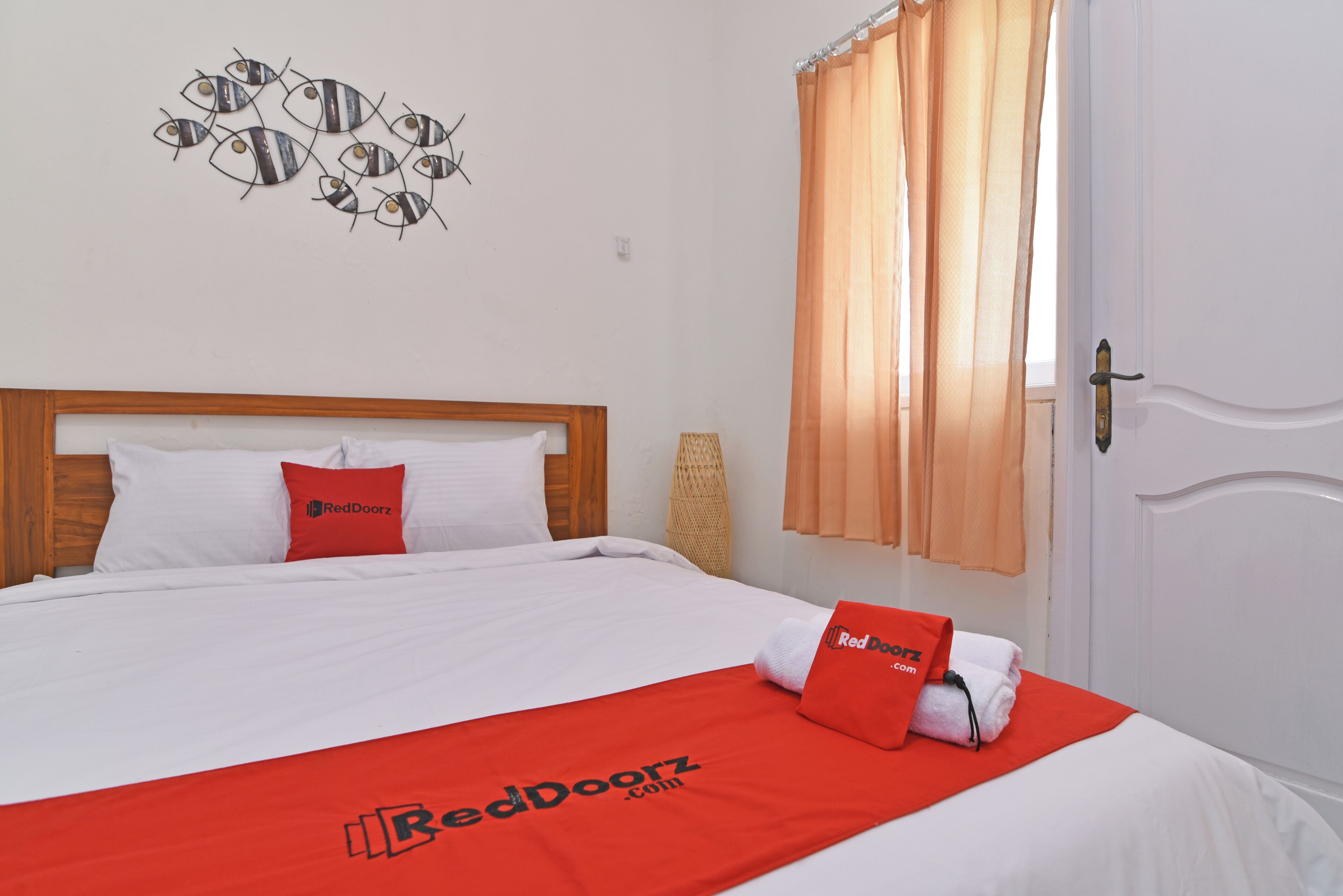 Una cama bien hecha con sábanas blancas y una manta roja, cabecera de madera, decoración de peces en la pared y una ventana con cortinas de encaje.