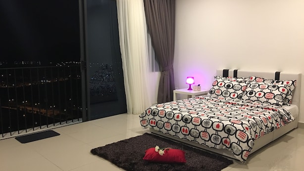 A bedroom with a large window, a bed with patterned bedding, a nightstand with a lamp, and a dark rug. - 1 Bedroom Apartment @ Trefoil Setia Alam (Shah Alam)