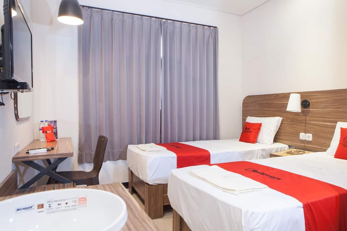 Habitación de hotel con dos camas individuales, un cabecero de madera, un escritorio con silla, una mesita pequeña, un espejo y una ventana con cortinas.
